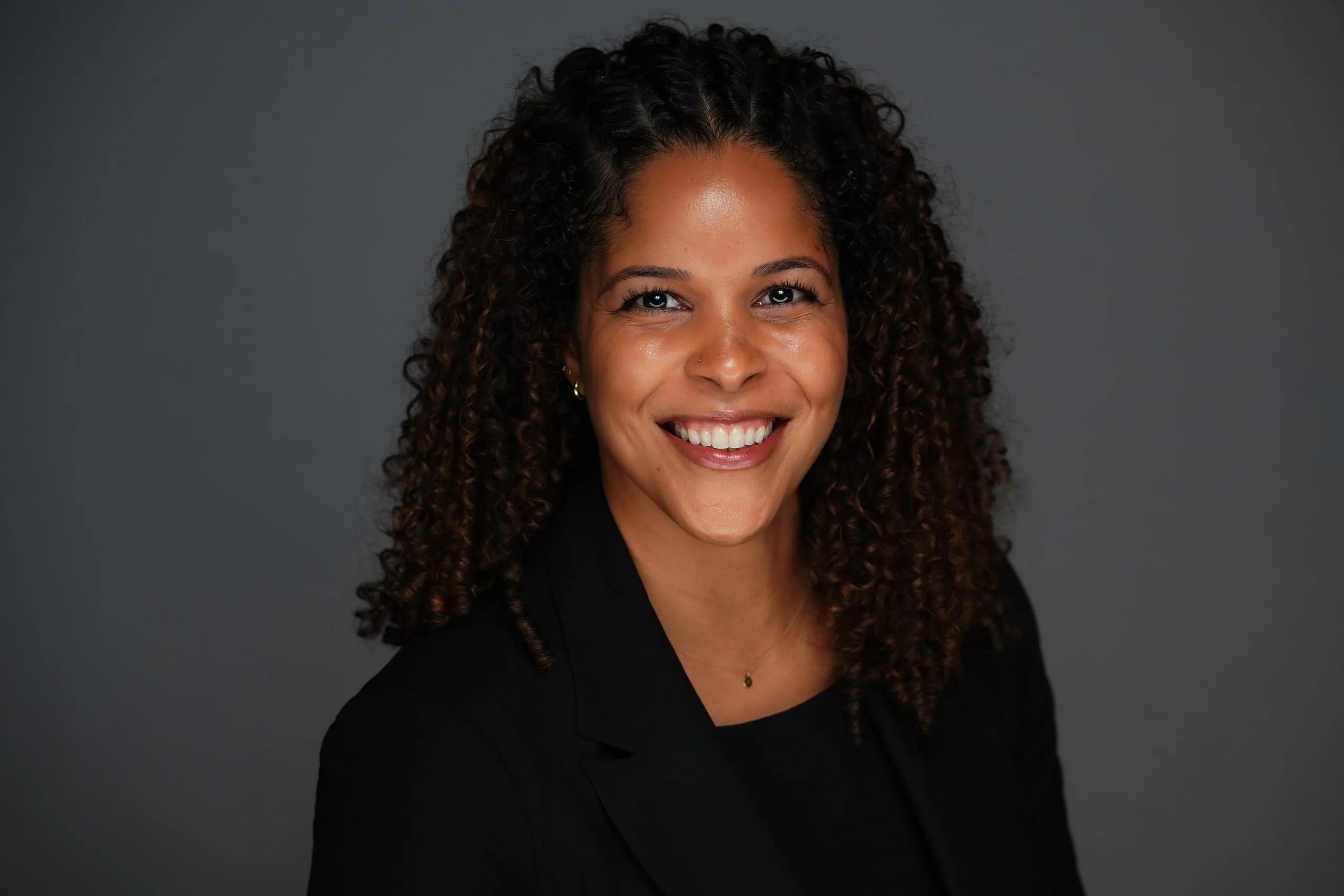 Marissa Gbenro smiling in a studio portrait, wearing a black blazer against a charcoal-grey backdrop.